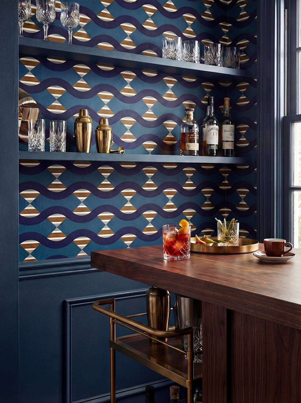 Bar setting with a wooden counter, drinks, and shelves with bottles and glasses against a decorative blue wall.